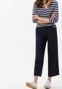 Pantalon large en lin bleu marine associé à un haut à manches longues rayé bleu marine et blanc avec un col en V. Des chaussures blanches à lacets complètent la tenue.