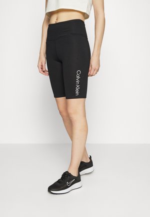 Femme portant un short de cycliste noir Calvin Klein et des baskets Nike noires, debout devant un fond blanc uni.