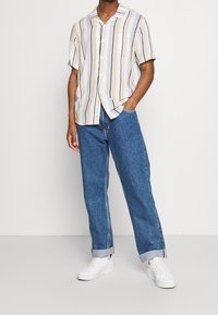 Camicetta a maniche corte a righe beige, crema e blu navy, abbinata a jeans in denim blu e sneakers bianche. La camicetta presenta una tasca e bottoni.