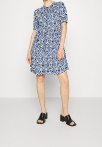 Vestido azul de manga corta con un patrón geométrico, cinturaCeñida y corte en A. Combinado con sandalias negras de tacón bloque abierto.