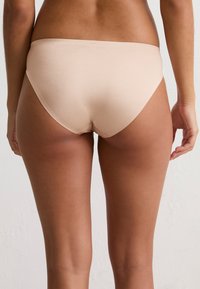 Vrouw in naadloze beige ondergoed, gezien van achteren tegen een eenvoudige witte achtergrond.