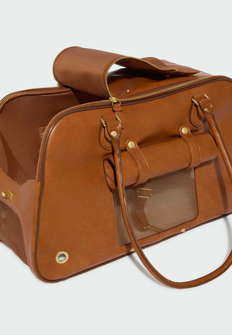 Trasportino per animali in pelle marrone con finiture dorate, finestra in rete, tracolle e zip superiore parzialmente aperta, su uno sfondo semplice e chiaro.