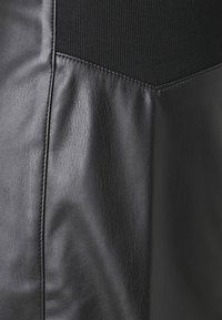 Svart läderplagg med en texturerad yta, som har en kontrasterande del i matt svart stickad material och synliga sömnadsdetaljer.