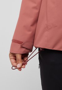 Roze buitenjasje met een gestructureerde stof, een verstelbare trekkoord aan de onderrand en een klittenbandsluiting bij de mouwen.