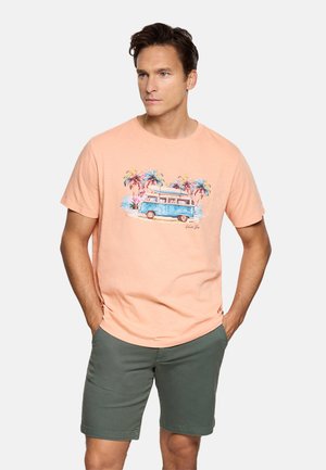 Hombre con camiseta color melocotón con gráfico de furgoneta azul y palmera, combinada con pantalones cortos verde oliva, de pie con las manos en los bolsillos.