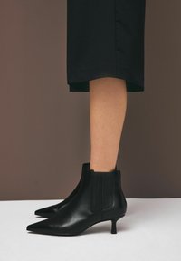 Černé kožené kotníkové boty s špičatou špičkou, vybavené elastickými bočními panely a nízkým jehlovým podpatkem. Hladká textura, minimalistický design.
