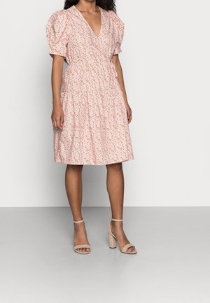 Gaun floral dengan lengan pendek menggelembung berwarna pink muda, pinggang yang terikat, dan hem yang melebar. Dipasangkan dengan sandal hak tinggi nude.