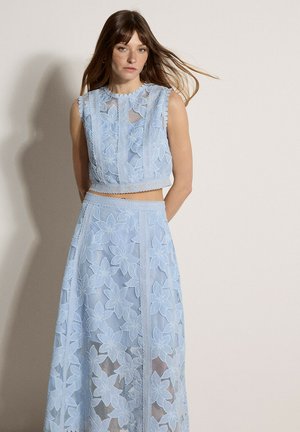 Kobieta ubrana w bezrękawowy, jasnoniebieski koronkowy top w kwiatowy wzór oraz pasującą długą spódnicę, stoi z rękami za plecami na tle jednolitej ściany.