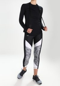 Svart långärmad träningströja med en lätt glans, kombinerad med svarta capri leggings med ett vitt och grått geometriskt mönster. Inkluderar sportskor.