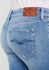 Jeans de mezclilla azul claro con una etiqueta de cuero que dice "REPLAY" y un sutil patrón de olas en el bolsillo trasero. Diseño clásico de cinco bolsillos.