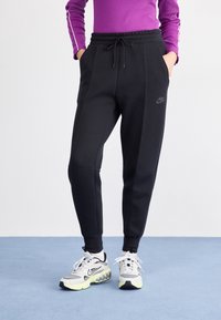 Person som bär svarta Nike-byxor och vita Nike-sneakers, står på ett blått golv med händerna i fickorna.