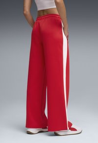 Red wide-leg sweatpants with white side stripes, elastic waistband, and pockets. Smooth fabric with a relaxed fit, paired with white shoes.
