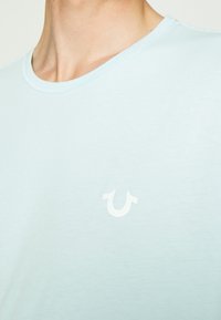 Světle modré bavlněné tričko s kulatým výstřihem a krátkými rukávy, s bílým logem na levé části hrudi. Měkká textura, minimalistický design.