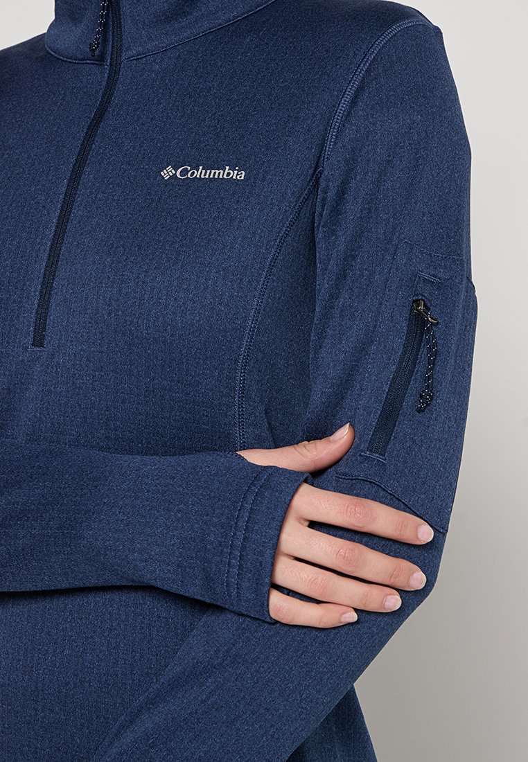 Primo piano di una persona che indossa un pullover a maniche lunghe blu Columbia con fori per i pollici e una tasca con zip sulla manica sinistra.