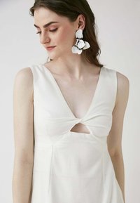 Weißes Kleid mit V-Ausschnitt, verdrehtem Vorderteil und Ausschnitt an der Brust. Das Model trägt große Blumenohringe. Glattes, untexturiertes Material.