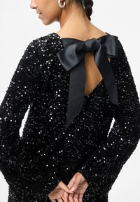 Kobieta w czarnej sukience z cekinami, z odkrytymi plecami, związanej dużą satynową wstążką, z dłońmi złożonymi za plecami.