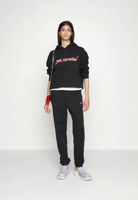 Hanorac negru cu text grafic, design crop, asortat cu pantaloni de trening negri. Modelul poartă pantofi slip-on gri deschis și are o geantă roșie.