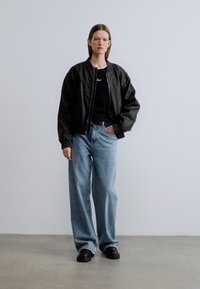 Veste bomber noire portée sur un t-shirt noir, associée à un jean à jambes larges de couleur claire et des chaussures noires. Texture lisse, coupe décontractée.
