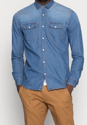 Mann trägt ein eng anliegendes, denimblaues Hemd mit Brusttaschen und beige Hosen, Hände in den Taschen, vor einem schlichten Hintergrund.