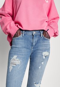 Jasnoniebieskie zniszczone dżinsy z poszarpanymi łatami i średnim stanem, zestawione z jasnoróżowym, oversize'owym swetrem.