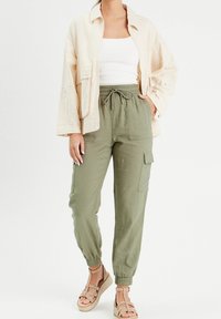 Veste beige clair, débardeur blanc côtelé et pantalon cargo vert olive avec poignets élastiques; associé à des sandales à brides couleur tan.