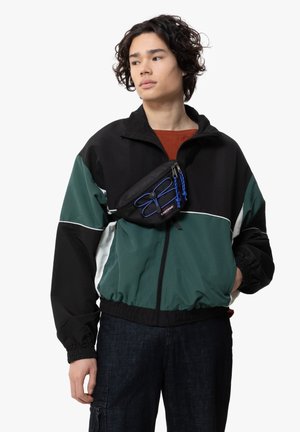 Veste coupe-vent noire et verte avec des accents blancs, dotée d'un zip sur le devant, de poignets élastiques et d'une petite banane noire avec des détails bleus.