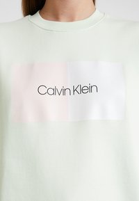 Světle zelená mikina s obdélníkovým obrázkem obsahujícím růžové a bílé části s textem "Calvin Klein" černě.