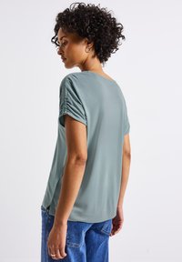 Haut vert clair à manches courtes avec une coupe décontractée, présentant des détails froncés aux épaules et une texture lisse. Associé à un jean bleu.