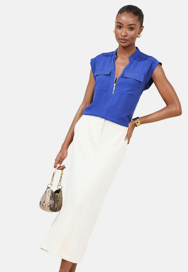 Blusa blu a maniche corte con collo a zip e due tasche frontali, abbinata a una gonna crema lunga fino alla caviglia. La modella tiene una borsa a tracolla con motivo.
