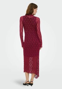 Robe en dentelle bordeaux à manches longues avec un motif ajouré, col montant et ourlet asymétrique, associée à des sandales noires à talons.