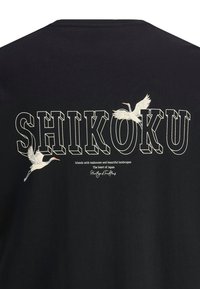 Melnas kokvilnas T-krekls ar vārdu "SHIKOKU" treknrakstā baltajā krāsā, apakšā ar divām grāmatu stārķu un tekstu par Japānu.