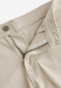 Beige pamut nadrág cipzárral és fémből készült gombos záródással. Varrott varratokkal és puha tapintással rendelkezik. Belseje egyszínű krémszínű béléssel van ellátva.