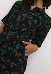 Combinaison noire à manches courtes avec motif botanique vert, dotée d'un col à revers, d'une fermeture à boutons et d'une taille cintrée avec un cordon de serrage.