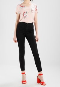 Blouse florale rose clair à manches courtes associée à un jean skinny noir taille haute et des sandales à talons blocs rouges. Textures de tissu lisses.