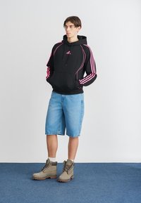 Hoodie preto com detalhes em rosa e logotipo, combinado com calções de ganga azul claro e botas cinzas. Apresenta bolso frontal e capuz com cordão.