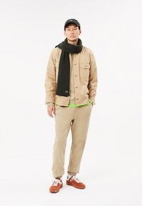 Giacca di cotone beige sovrapposta a una felpa verde, abbinata a pantaloni tapered beige, una sciarpa a coste scura e sneakers in suede marroni. Cappellino nero.