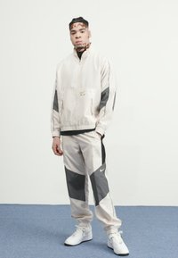 Light beige windbreaker with gray accents, paired with matching joggers. Features a zip front, elastic cuffs, and Nike logo. White sneakers.