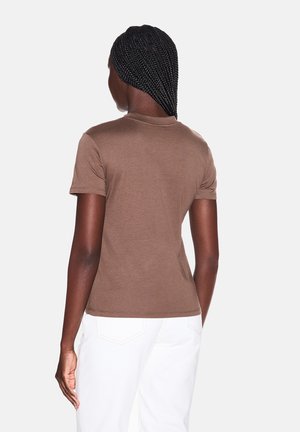 Persona con i capelli intrecciati che indossa una maglietta marrone a maniche corte e pantaloni bianchi, in piedi di spalle su uno sfondo semplice.