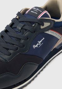 Σκούρο μπλε αθλητικό παπούτσι με υλικά από συνθετικό κα�ι δέρμα με υφή, διαθέτει αντίθετες ανοιχτές λεπτομέρειες και λογότυπο στο πλάι. Κοινοί κορδόνια και επένδυση στη γλώσσα.