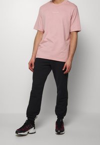 Vaaleanpunainen puuvillapaitainen t-paita, jossa on valkoinen logo, yhdistettynä mustiin kapeutuviin collegehousuihin, joissa on sivuraitakangas, ja monivärisiin urheilukenkäisiin.