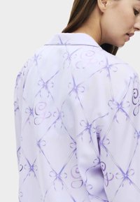 Hellblaues Seidenhemd mit einem Diamantmuster, Bows und geschwungenen Buchstaben. Kragen-Design mit glatter Textur und subtiler Ausstrahlung.