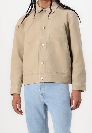 Personne portant une veste beige boutonnée avec grandes poches avant et un jean bleu clair sur un fond blanc uni.