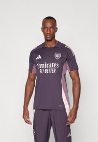 Maillot de football Arsenal en violet foncé, avec des accents blancs, col rond, manches courtes, et le logo du sponsor "Emirates Fly Better" sur le devant.