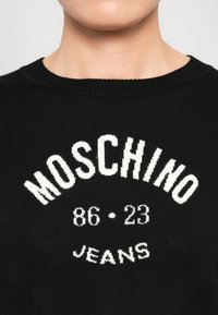Čierny sveter s bielym pleteným textom "MOSCHINO 86 • 23 JEANS" na hrudi, nosený osobou zobrazenou od krku po hornú časť hrude.
