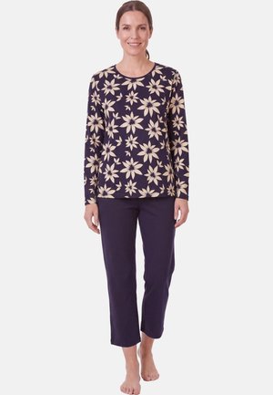 Kvinde smilende iført en marineblå bluse med lange ærmer med beige blomsterprint og ensfarvede marineblå kortbukser, barfodet på hvid baggrund.