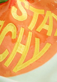 Vyšívaná tkanina s výraznými žltými písmenami na oranžovom pozadí s jemnou textúrou, efektom miešaného prania a žiarivou farbou.