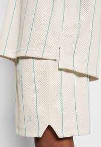 Shorts en maille beige avec des rayures verticales bleu sarcelle et le texte "Air Jordan", portés par une personne, montrant des détails d'ouverture latérale et un tissu perforé.