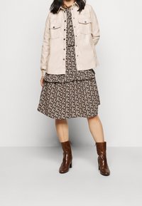 Beigefärgad läderjacka med två bröstfickor över en svart blommig klänning med lager på lager volanger. Bruna klackade anklarstövlar med textur.