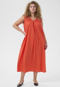 Langes, orangefarbenes, ärmelloses Kleid mit gerafftem Ausschnitt, lockerer Passform und Eingrifftaschen. Glatte Textur, knielang, kombiniert mit schwarzen Schuhen.