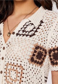 Detailní záběr na krémový háčkovaný svetr s hnědými a béžovými čtvercovými vzory, dřevěnými knoflíky a zlatým náhrdelníkem s přívěskem na ženě.